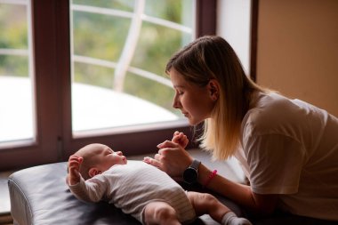 Sevgi dolu anne yeni doğmuş bebeğine bakıyor, pencerenin yanındaki minicik ellerini nazikçe tutuyor, sakin bir gündüz saatinde yumuşak, doğal ışıkta güneşleniyor.