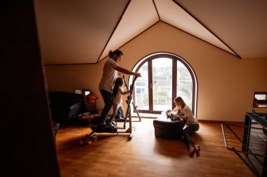 Anne ve baba, oturma odasında yeni doğmuş bebeklerine bakarken eliptik bir makinede egzersiz yapıyor. Evde canlı ve sağlıklı bir aile ortamı yaratıyor.