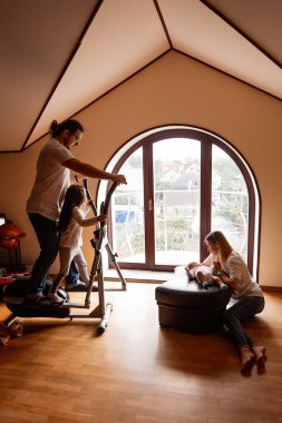 Baba ve kızı, anne yeni doğmuş bebeklerine bakarken eliptik bir eğitmen üzerinde egzersiz yapıyor, rahat oturma odalarında doğal ışıkla dolu neşeli bir atmosfer yaratıyor.