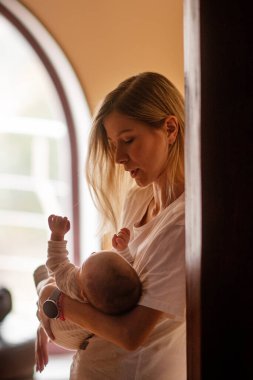 Genç anne, yeni doğmuş bebeğini bir pencerenin yumuşak parıltısıyla kucaklıyor. Sevgi dolu, bağ dolu ve huzurlu bir ev ortamında sıcaklığı besleyen hassas bir anı yakalıyor.