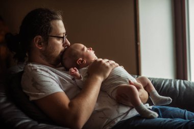 Yeni doğmuş bebeğini öpen genç bir baba pencerenin yanındaki rahat bir kanepede otururken ebeveynliğin neşesine değer veriyor ve şefkat ve şefkat ile dolu derin, sevgi dolu bir bağ besliyor.