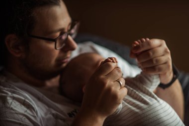 Baba, yeni doğmuş bebeğinin minik elini nazikçe tutarak yumuşak, loş bir ortamda sevgi, bağ ve ebeveyn bağı yaratıyor.