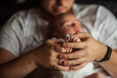 Baba, uyuyan yeni doğan bebeğinin minik elini tutuyor, sevgi, ilgi ve korunma dolu sıcak ve samimi bir anda.