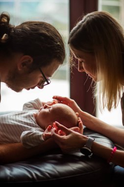 Genç anne ve baba, kanepede dinlenen yeni doğmuş bebeklerine sevgi dolu bir şekilde bakıyorlar. Küçük ellerini nazikçe sıcacık ve sıcak bir oturma odasında tutuyorlar.