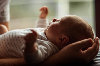Yeni doğan bebek, bir yetişkinin kollarında huzur içinde yatar ve hassas bir ilgi ve bağlılık anı yakalar. Yumuşak ışıklandırma bebeğin hassas özelliklerini ve sakin ifadesini vurgular.