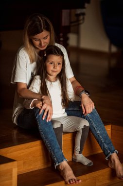 Sevecen anne, genç kızını sevgiyle kucaklıyor ve birlikte tahta basamaklarda oturuyorlar, sessiz ve samimi bir anın tadını çıkarıyorlar. İçerideki sıcak ortam bağlantılarını güçlendiriyor.