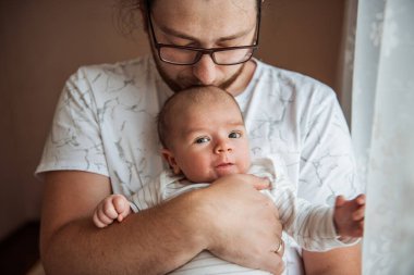 Bir babanın yeni doğmuş bebeğini pencerenin yanında kucakladığı o hassas an. Yumuşak ışıklandırma, ebeveynliğin özünü yakalayarak, aralarında paylaşılan bağın ve sıcaklığın altını çiziyor.