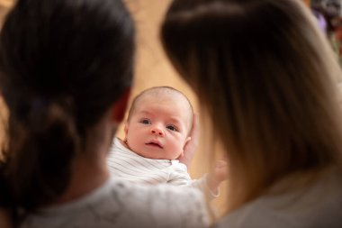 İki ebeveyn yeni doğan bebeklerini kucağında tutuyor, ona derin bir sevgi ve özenle hayranlık duyuyorlar, neşe, şefkat ve aile bağlarının güzelliği ile dolu doyurucu bir an yaratıyorlar.