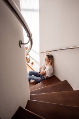 Genç anne, yeni doğmuş bebeğini emzirirken evdeki ahşap merdivenlerde rahatça oturur, anneliğin ve sevgi ve özenle dolu beslenme anlarının tadını çıkarır.