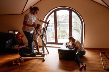 Anne, büyük bir pencereden gelen ışıkla dolu parlak bir oturma odasında yeni doğan bebeklerine bakarken baba ve kız birlikte eliptik bir makinede egzersiz yapıyorlar.