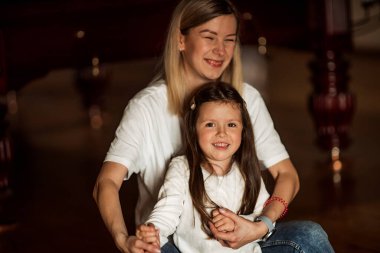 Anne ve kız oturuyor ve el ele tutuşuyor, ikisi de beyaz tişörtler giyiyor ve loş ışıklı bir odada gülümsüyor, arka planda ahşap bir zemin ve kırmızı kolonlar var.