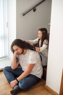 Genç kız, babasının uzun saçlarını, o merdivenlerde otururken şakacı bir şekilde örüyor. Sahne, evde birlikte vakit geçirmenin verdiği mutluluk ve bağın vurgulandığı hassas bir aile anını yakalıyor.