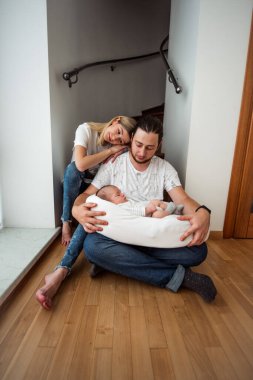 Genç anne ve baba, merdivenlerin altında oturmuş, yeni doğmuş bebeklerini rahat bir bebek yuvasında kucaklıyorlar. Huzurlu evlerinde sevgi ve şefkati somutlaştırıyorlar.