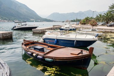 Geleneksel ahşap tekne, arka planda Karadağ 'ın pitoresk dağlarıyla Kotor Körfezi' nin sakin sularında modern motorbotlarla birlikte demirlemiş durumda.