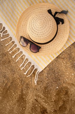 Plaj havlusu, hasır şapka ve kumların üzerinde duran güneş gözlükleri plajda rahatlatıcı bir yaz günü izlenimini uyandırıyor. Seyircileri tasasız bir tatil hayal etmeye davet ediyor.