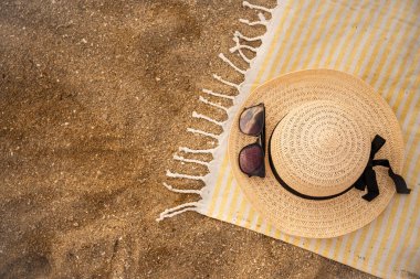 Sıcak kumda dinlenen sarı çizgili plaj havlusu, hasır şapka ve güneş gözlüğü eşliğinde, okyanus tarafından rahatlatıcı bir yaz günü için mükemmel bir ortam yaratıyor.