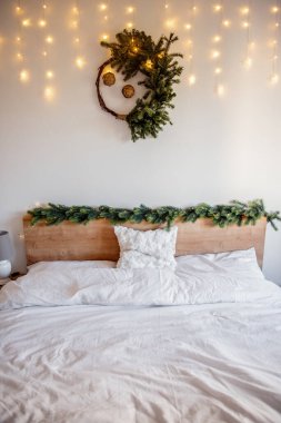 Yatağın üzerinde asılı duran Noel çelengi yatak odasını köknar dallarıyla aydınlatıyor yatak başlığını, beyaz yastığı ve battaniyeyi süsleyerek rahat ve şenlikli bir ortam yaratıyor.
