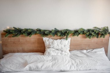 Beyaz çarşaflar ve beyaz yastıkla döşenmiş bir yatağın ahşap yatak başlığını süsleyen küçük ışıklı Noel çelengi rahat ve şenlikli bir atmosfer yaratıyor.