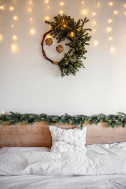 Noel çelengi, yatağın üzerinde çam kozalaklarıyla süslenmiş. Peri ışıkları, rahat bir yatak odasını hafifçe aydınlatıyor. Tatil sezonu için sıcak ve şenlikli bir atmosfer yaratıyor. Şenlikli iç mekan 