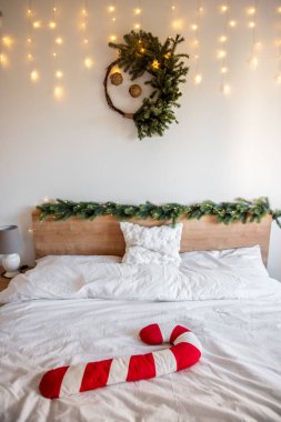 Noel süslemeleri peri ışıkları ve köknar dalları ile bir yatakta dinlenen şeker kamışı yastığı ve duvarda asılı köknar çelengi, neşeli ve rahat bir atmosfer yaratır.
