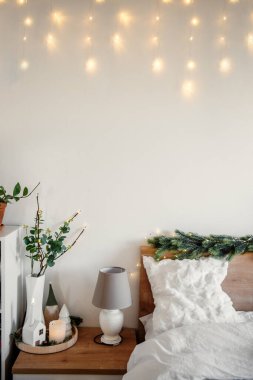 Noel ışıkları beyaz bir duvara asılıyor ve yatak başlığını beyaz yastıklar ve battaniyelerle süslüyor. İçinde lamba olan bir komodin. Bitkiler ve Noel süsleri. Sıcak bir atmosfer yaratıyor