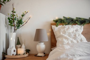 Noel ışıkları köknar dallarını, mumları ve dekoratif evleri aydınlatıyor. Beyaz çarşaflı yatak odasında rahat bir atmosfer yaratıyor.