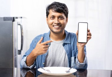 Hintli bir etnik erkek beyaz ekranı işaret ediyor. Gülümseyen bir yüzü var. Yemek masasının yanında oturuyor ve evde boş bir tabak var.