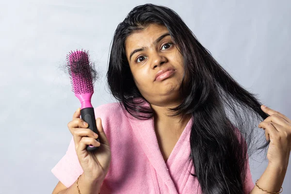A worried Indian woman in bathrobe facing hair fall problem looks at camera with hair brush on white background