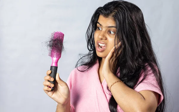A worried Indian woman in bathrobe facing hair fall problem looks at her hair brush with tangled hair on white background