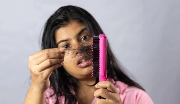 Selective focus on a pink hair brush full of fallen hair held in hand by a worried Indian woman on white background