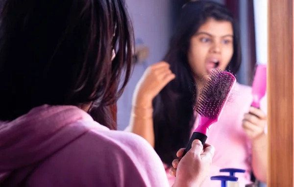Selective focus on a pink hair brush full of fallen hair held in hand by a worried Indian woman in front of mirror