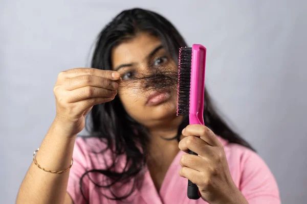 Selective focus on a pink hair brush full of fallen hair held in hand by a worried Indian woman on white background