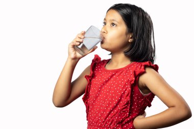 Kırmızı elbiseli Hintli bir kız çocuğu beyaz arka plandaki bardaktan su içiyor.