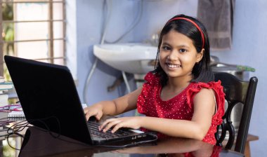 Kırmızı elbiseli şirin bir Hintli kız evde dizüstü bilgisayarla ders çalışıyor.