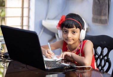 Kırmızı elbiseli şirin bir Hintli kız evde bilgisayar ve kulaklıkla ders çalışıyor.