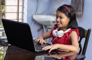 Kırmızı elbiseli şirin bir Hintli kız evde bilgisayar ve kulaklıkla ders çalışıyor.