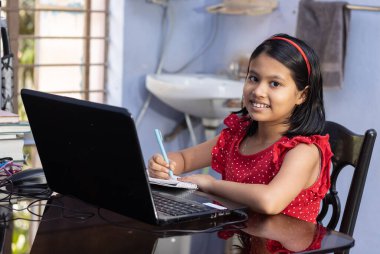 Kırmızı elbiseli şirin bir Hintli kız evde dizüstü bilgisayarla ders çalışıyor.