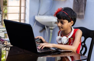 Kırmızı elbiseli şirin bir Hintli kız evde bilgisayar ve kulaklıkla ders çalışıyor.