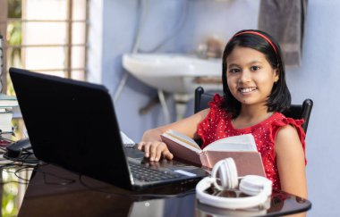 Kırmızı elbiseli şirin bir Hintli kız evde bilgisayar ve kulaklıkla ders çalışıyor.