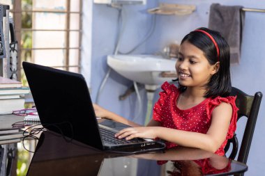 Kırmızı elbiseli şirin bir Hintli kız evde dizüstü bilgisayarla ders çalışıyor.