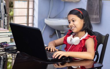 Kırmızı elbiseli şirin bir Hintli kız evde bilgisayar ve kulaklıkla ders çalışıyor.
