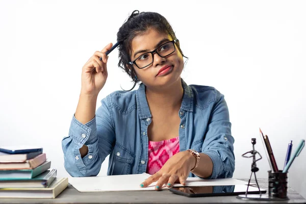 A pretty young Indian college student in eyeglasses thinking while studying on study table and white background
