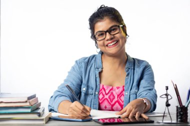 Gözlüklü güzel bir Hintli üniversite öğrencisi çalışma masasında ve beyaz arka planda okuyor ve yazıyor.