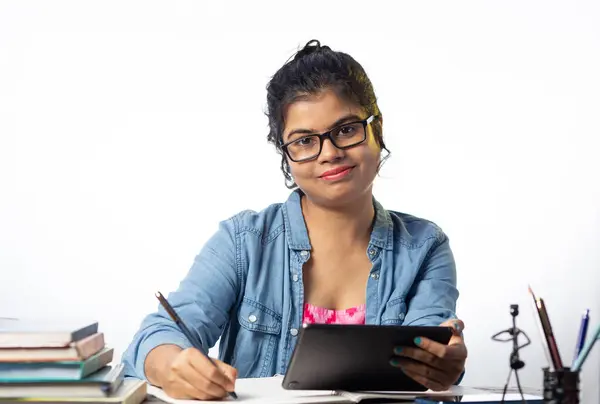 A pretty young Indian college student in eyeglasses studying with tablet on study table and white background