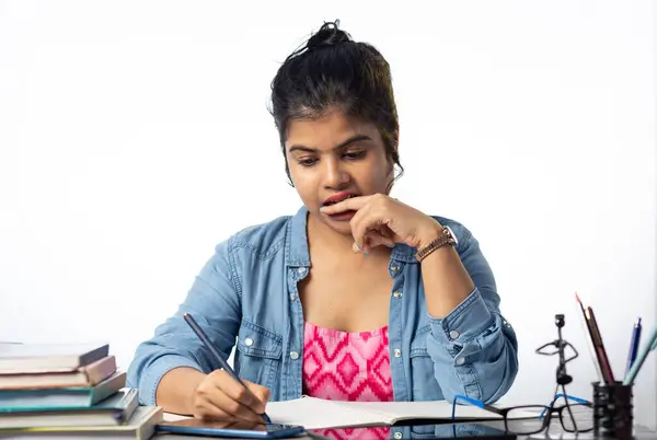 A pretty young Indian college student studying and biting finger on study table and white background