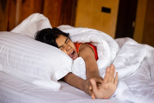 A young Indian pretty woman waking up and yawning on a cozy white bed and pillow covering blanket