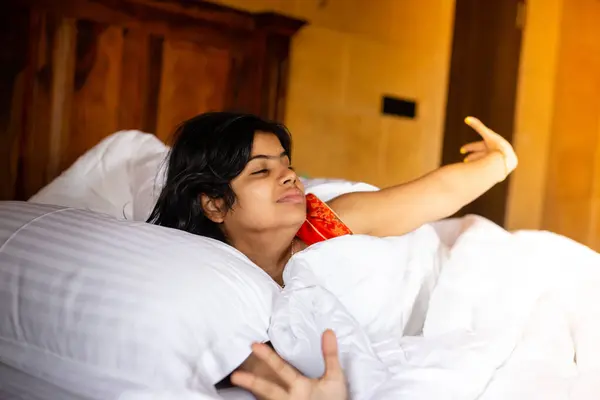 A young Indian pretty woman waking up and yawning in the morning on a cozy white bed and pillow covering blanket