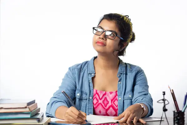 A pretty young Indian college student in eyeglasses studying and looking sideways on study table and white background