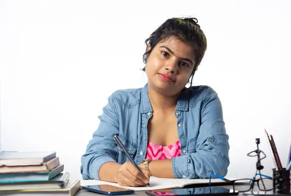 A pretty young Indian college student studying and looking at camera on study table and white background