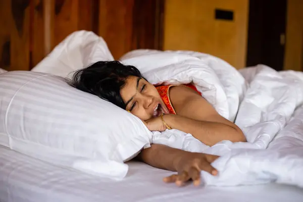 A young Indian pretty woman waking up and yawning on a cozy white bed and pillow covering blanket